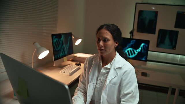 Dedicated female geneticist working at her desk during the night. The researcher is analyzing dna data on her computer in a modern lab