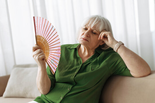 Senior woman cooling herself with handheld fan at home living room. Mature caucasian female feeling hot and uncomfortable, illustrating summer heat or menopause symptoms in retirement lifestyle
