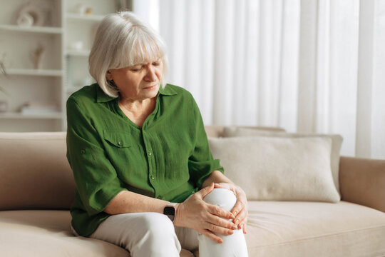 Senior woman holding painful knee while sitting on sofa at home. Mature caucasian female experiencing joint pain or arthritis, illustrating aging health problem in retirement lifestyle
