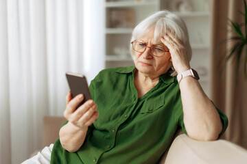Senior woman with glasses looking at smartphone with worried expression while sitting at home. Mature caucasian female reading message or news online on mobile device © Anastasiya