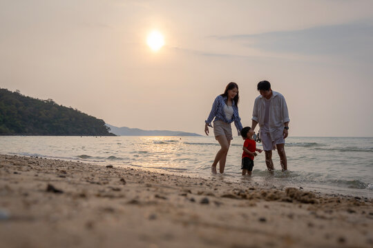 Happy Asian family with a young son enjoying sunset on the beach, playing in the sand and walking together during summer vacation.