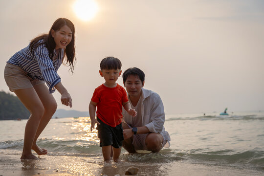 Happy Asian family with a young son enjoying sunset on the beach, playing in the sand and walking together during summer vacation.