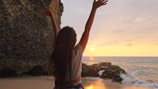 Carefree young woman stands on a tropical beach at sunset with arms raised in celebration. Long dreadlocks, flowing maxi skirt, and cropped top highlight summer travel, wellness, and lifestyle concept