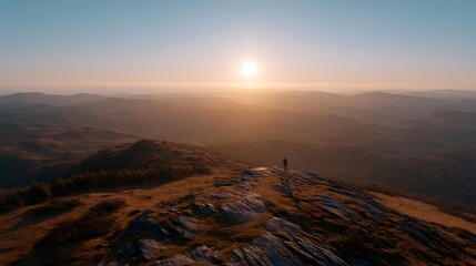 A solitary figure stands on a mountain summit at sunrise overlooking a vast hazy landscape bathed in golden light