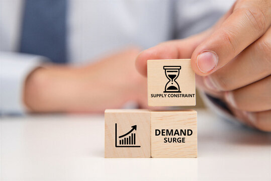 Hand placing wooden block with supply constraint hourglass symbol near demand surge and graph blocks signifying business challenges