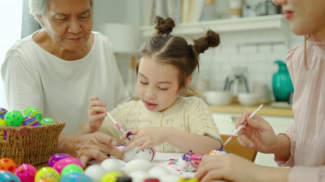 Happy multi-generation family with grandmother, mother, and daughter painting colorful Easter eggs in a bright kitchen. A joyful holiday tradition and creative activity for kids at home.