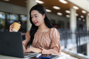 Fototapeta premium Asian young adult woman freelancer works laptop computer holding coffee cup modern cafe casual professional remote work business education student online
