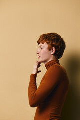 Profile portrait of redhead young man wearing brown turtleneck sweater standing against beige background with thoughtful expression and hand near chin. © SHOTPRIME STUDIO