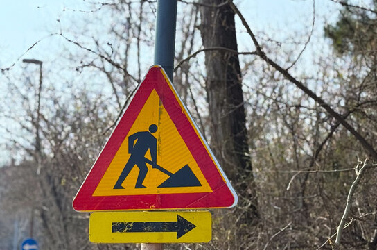 Roadwork warning sign with arrow direction placed near forest roadside. Traffic regulation, infrastructure maintenance, driver attention, safety awareness and transportation management concept.