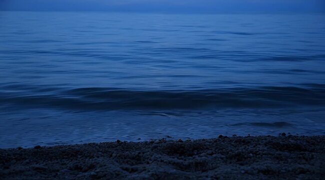 Calm, dark blue water gently laps against a pebble-strewn shore under a twilight sky