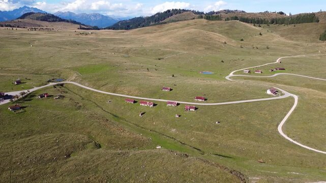 Aerial drone footage revealing the Marcesina plateau, a high alpine plain on the Asiago Plateau, showcasing rows of vintage &ldquo;casoni&rdquo; with rusted roofs, expansive green pastures and meandering mountain