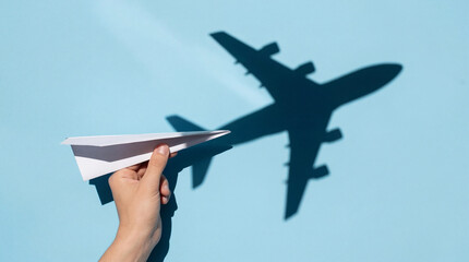 Hand holds a white paper airplane casting a shadow of a large airplane symbolizing the start of a journey from a small idea