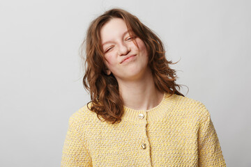 Young woman with curly brown hair wearing yellow knitted sweater makes funny face with closed eyes...