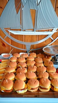 Rows of mini cheeseburger sliders on wooden catering tray. Many identical mini burgers with toothpicks served on a wooden board against a nautical wall decoration.