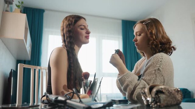 Caucasian Duo Testing Perfume And Makeup At Dining Table, Bright Window Background, One Holding Spray Bottle While Other Breathes In Scent, Playful Smiles, Makeup Tools Scattered On Glass Surface,