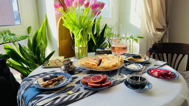Luxury spring brunch celebrating Maslenitsa and March 8th. Festive table with pink Prosecco, black caviar, Perm ham, and crepes. Elegant holiday morning with purple tulips on ikat tablecloth.