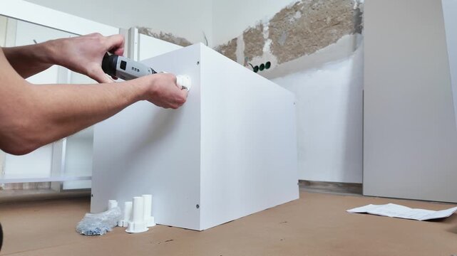 Wide Angle Contractor Securing Cabinet To Wall Caucasian Installer Using Power Driver To Fasten Brackets And Anchors, Reinforced Alignment, Protective Cardboard On Floor, Hardware Pile Close By