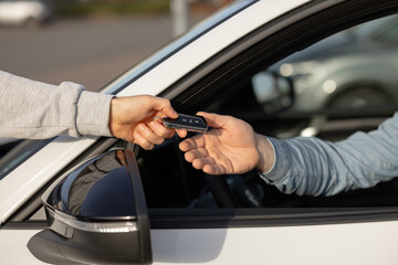 Hand of a person in gray sweatshirt passing car keys to another person inside a white vehicle, with blurred cars in the background in a parking area © Kateryna Muzhevska