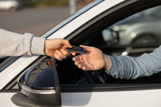 Hand of a person in gray sweatshirt passing car keys to another person inside a white vehicle, with blurred cars in the background in a parking area
