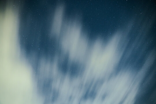 Long exposure night sky with drifting clouds forming soft motion trails against a deep blue background filled with distant stars, creating a dreamy and atmospheric celestial scene.
