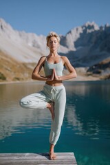 Fototapeta premium Female caucasian adult woman meditating yoga on a wooden dock by an alpine mountain lake at sunrise serene mindfulness balance