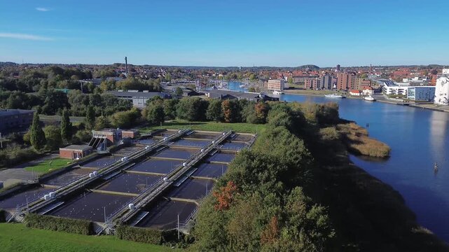 Danish wastewater treatment plant located near the waterfront, with clear views of Haderslev Fjord and the surrounding city. The facility includes large treatment basins
