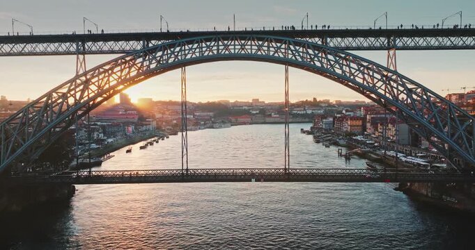 Dom Luis I Bridge steel arch structure connecting Porto and Vila Nova de Gaia, spanning Douro River with boats, showcasing the illuminated cityscape during a golden hour sunset. Aerial drone flight