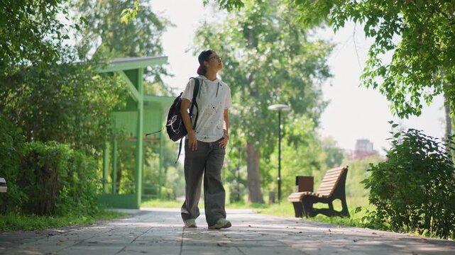 Strolling man under leafy shade, Person leisurely walking past greenery with backpack on warm afternoon, Individual taking slow walk in park with trees and backpack on sunny summer day