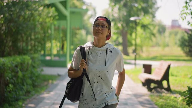 young adult walking sunlit park path lined with benches and gazebo carrying black backpack smiling in casual hoodie warm summer light relaxed urban stroll candid shallow depth portrait