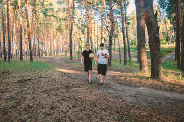 Fototapeta premium Two Teenage Friends Walking through a Sunny Pine Forest with a Soccer Ball and Smartphone during Golden Hour