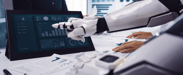 A robot collaborates with a human team in a modern office setting, analyzing data on a touchscreen display, showcasing technology's role in business productivity.Noogenesis