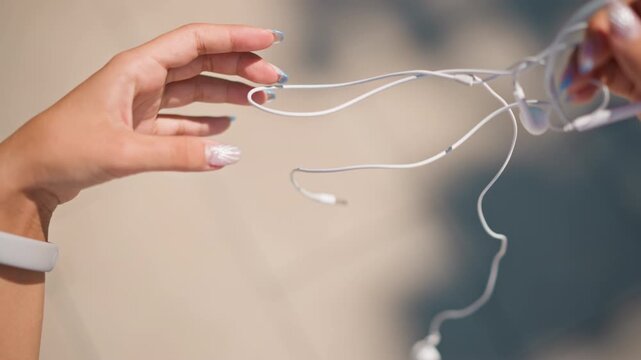 female hands untangling white earbuds on sunlit pavement, closeup of manicured fingers working through knots, wristwatch visible, soft shadows, patient methodical motions, commuting prep and everyday
