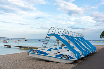 Obraz premium Row of blue and white pedal boats with water slides lined up on pebble beach ready for summer rental with calm sea. Beach water entertainment, family fun and active seaside holiday.