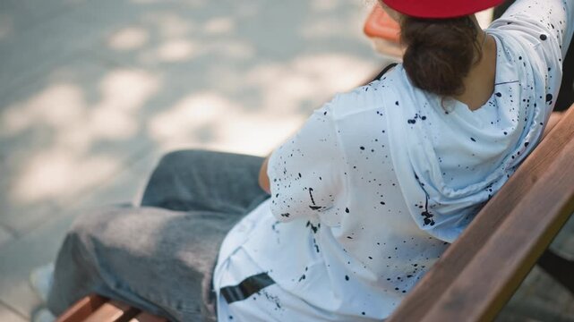 closeup cap and hands on bench, white speckled shirt, red visor, ponytail and sunglasses perched on cap, dappled shadows from trees, watch on wrist, relaxed posture, lunch container at side, quiet
