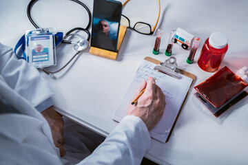Doctor writing notes on clipboard during online consultation with patient on smartphone