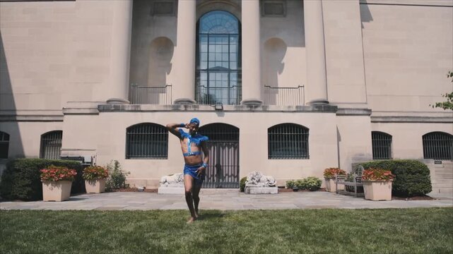 Black man in a blue costume voguing in a park in front of a building