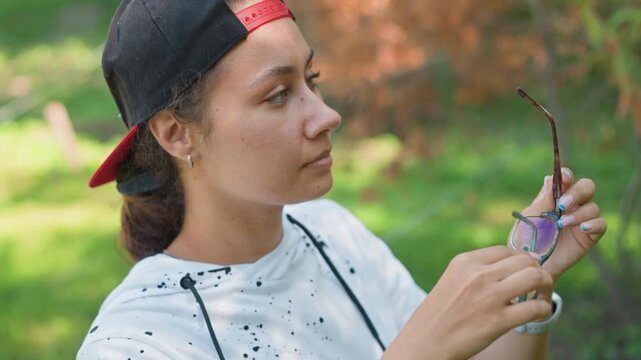 young woman checking sunglasses outdoors cap backwards attentive expression adjusting frames wristwatch detail leafy background dappled light closeups candid angles observational mood
