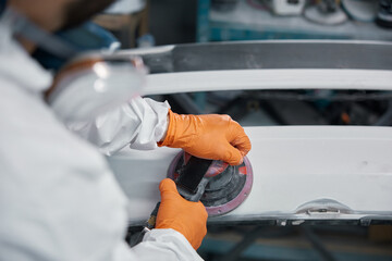 The technician is sanding the car hood in the workshop, wearing safety gear for repainting