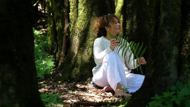Thoughtful mature woman sitting on forest floor enjoying quiet connection with nature. Peaceful female experiencing freedom, self reflection and personal wellbeing while spending mindful time alone in