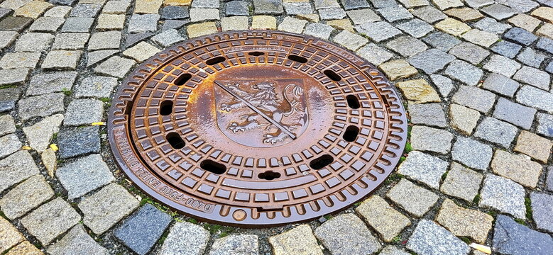 Ein gusseiserner, runder Kanaldeckel mit dem Stadtwappen von Herzogenaurach, eingebettet in ein klassisches Kopfsteinpflaster aus Granitsteinen. Ein St&uuml;ck Stadtgeschichte unter den F&uuml;&szlig;en.