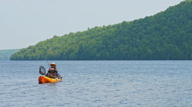 Woman casting and reeling while floating on kayak boat. Kayaking lake with scenic landscape.  Woman angler in a sun hat on motorized kayak enjoying peaceful summer vacation sunny day, active outdoor.