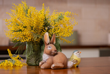 Easter bunny figurine with tit bird and mimosa bouquet, spring holiday decoration in the modern kitchen interior. Cozy spring composition good for Easter celebration and seasonal decor. © Hanna Aibetova
