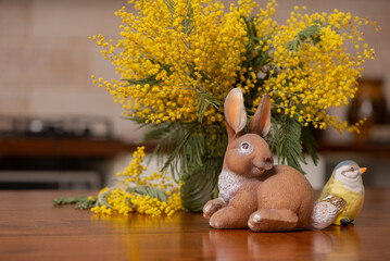 Easter bunny figurine with tit bird and mimosa bouquet, spring holiday decoration in the modern kitchen interior. Cozy spring composition good for Easter celebration and seasonal decor. © Hanna Aibetova
