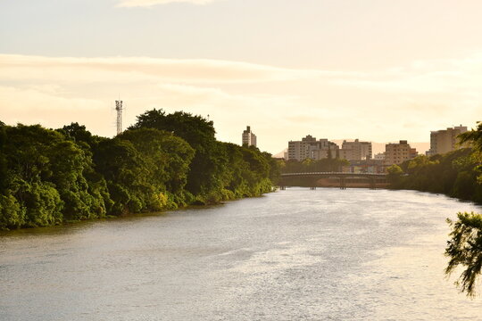 Paisagem fluvial urbana ao p&ocirc;r do sol com mata ciliar e silhueta de pr&eacute;dios