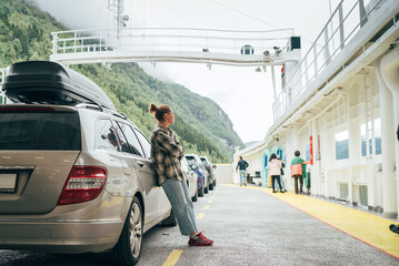 Woman leans against her car on Norwegian ferry surrounded by other vehicles with misty green mountains in background. Scene captures essence of relaxed travel, exploration in scenic Norwegian fjords