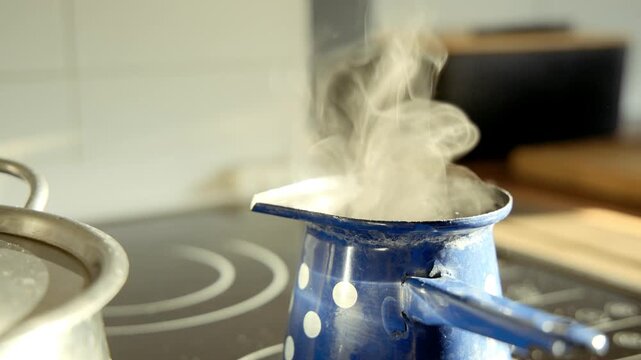 Sunlit kitchen scene shows a small blue enamel coffee pot with white polka dots steaming on a wooden board. Ceramic mugs and a kettle sit nearby, capturing a warm, cozy morning coffee moment at home.