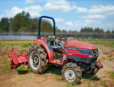chiba, katori - mar 26 2025: A red Japanese Mitsubishi MT240 tractor parked in a farm field at THE FARM resort, with a rotary tiller attachment and a small pot of bright flowers on the ground nearby.