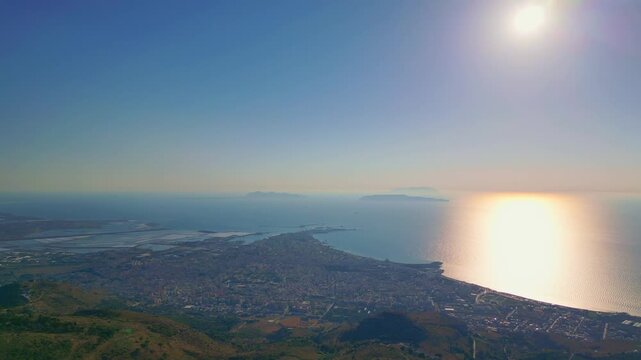 Cinematic aerial panorama of Trapani, Sicily emerging behind a hill. Distant coastal city skyline with the Mediterranean Sea in the foreground, dramatic landscape reveal and scenic travel view

