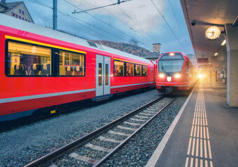 Red modern trains on mountain railway station in Swiss Alps in autumn at sunset. Speed trains with lights, railroad, railway platform in fall at dusk in St. Moritz, Switzerland. Bernina Express