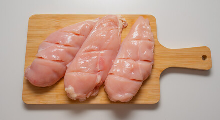 Three pieces of fresh raw chicken breast with light incisions placed on a wooden cutting board. Top view of healthy protein-rich poultry meat on a white background